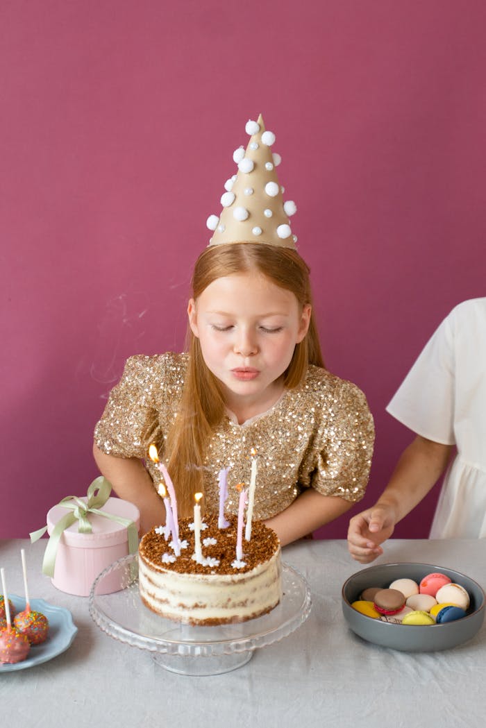 A child in a festive hat blows out candles on a birthday cake during a party.